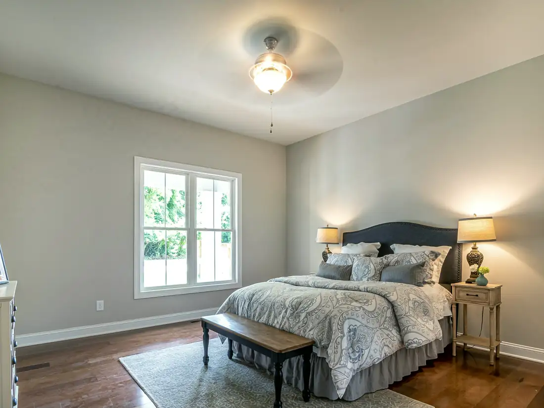 A bedroom with grey walls, a navy upholstered headboard, two bedside lamps, a wooden bench and a ceiling fan.