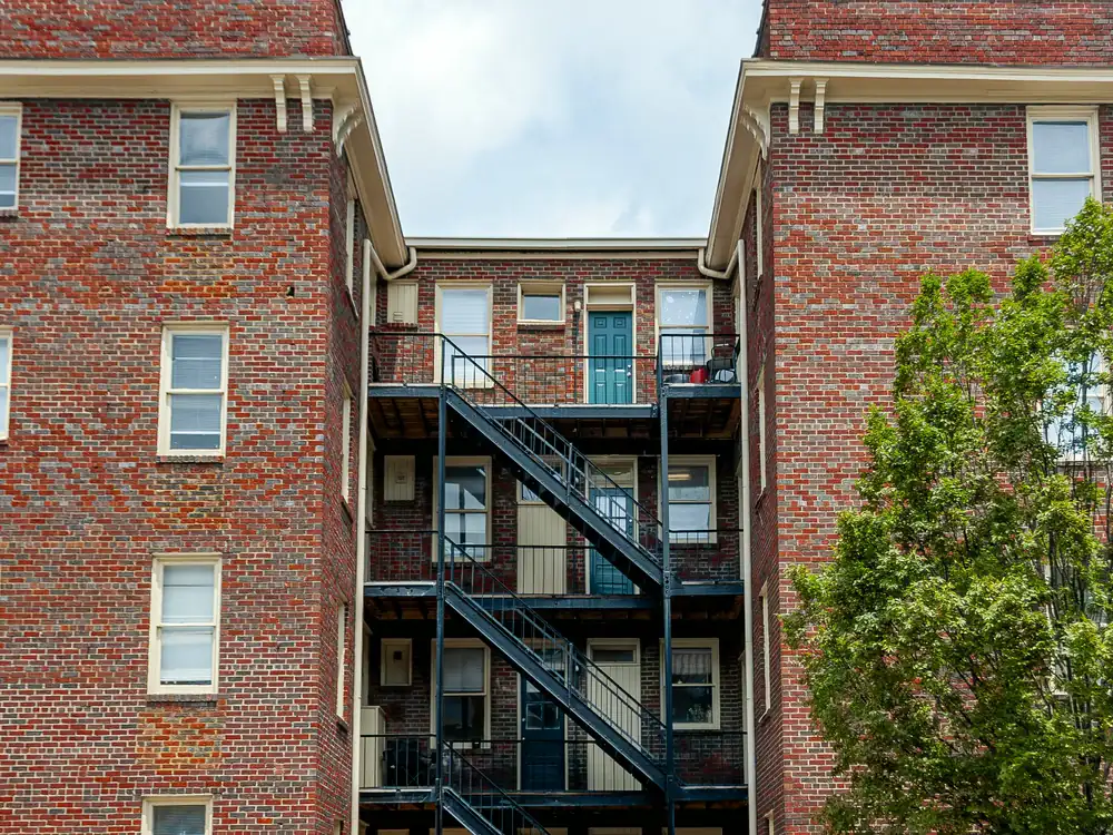 A four-storey brick apartment building with a black fire escape between two wings, against a bright sky.