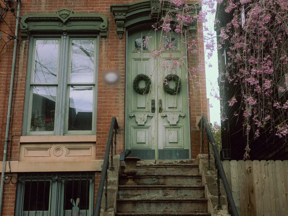 A narrow brick rowhouse with sage green double doors, carved window heads and a steep stone stoop, with a flowering tree beside it.