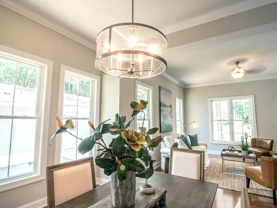 A dining table under a drum chandelier, open to a living area with a white sofa and two leather armchairs, grey walls and white windows.