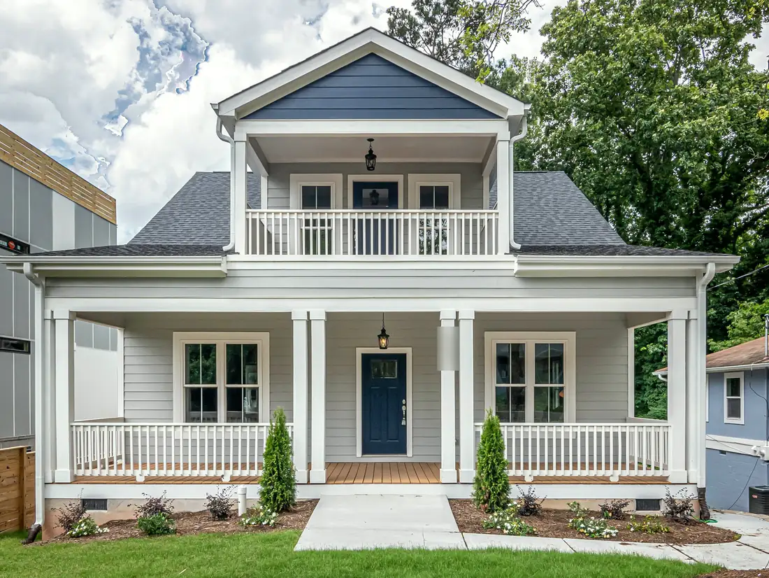 A grey two-storey house with a navy front door, white porch columns and a covered balcony porch above, behind a small planted front yard.