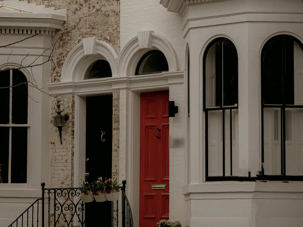 A painted white rowhouse with arched windows, a bay front and a red door at the top of a short iron-railed stoop.