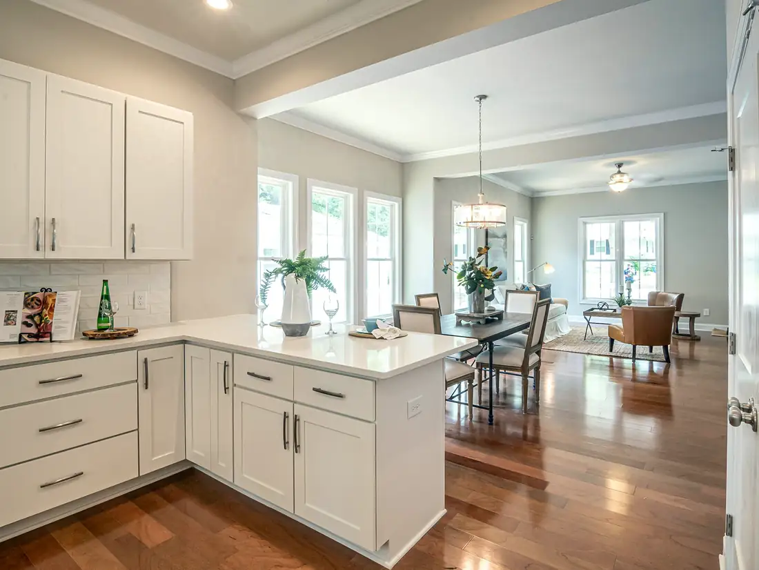 A white Shaker kitchen with quartz counters looking through to the dining table and living room on dark hardwood floors.